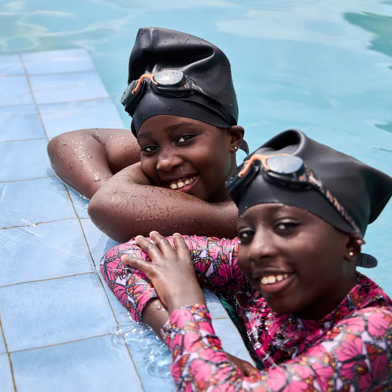 Two young students wearing swim caps and goggles smiling while leaning on the edge of a swimming pool.