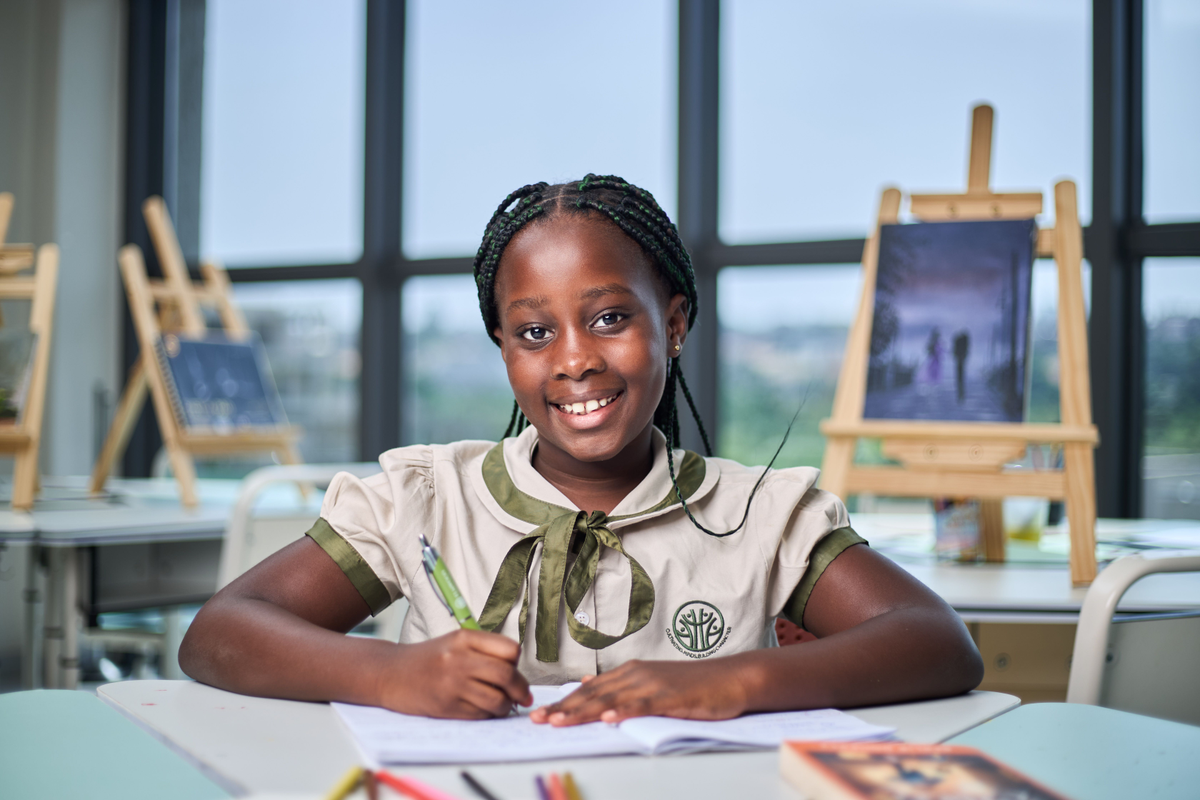 A smiling Elmwood International School student with braided hair sitting at a desk and writing in a notebook, with art easels in the background.