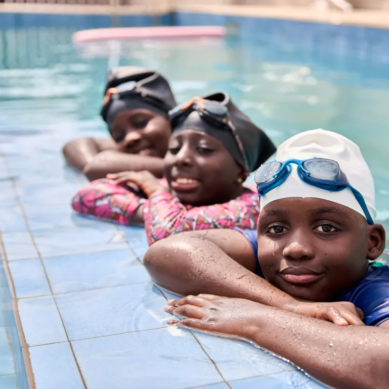 A young Elmwood International School student wearing a black chef's hat whisking ingredients in a large metal bowl with a smudge of flour on her forehead.