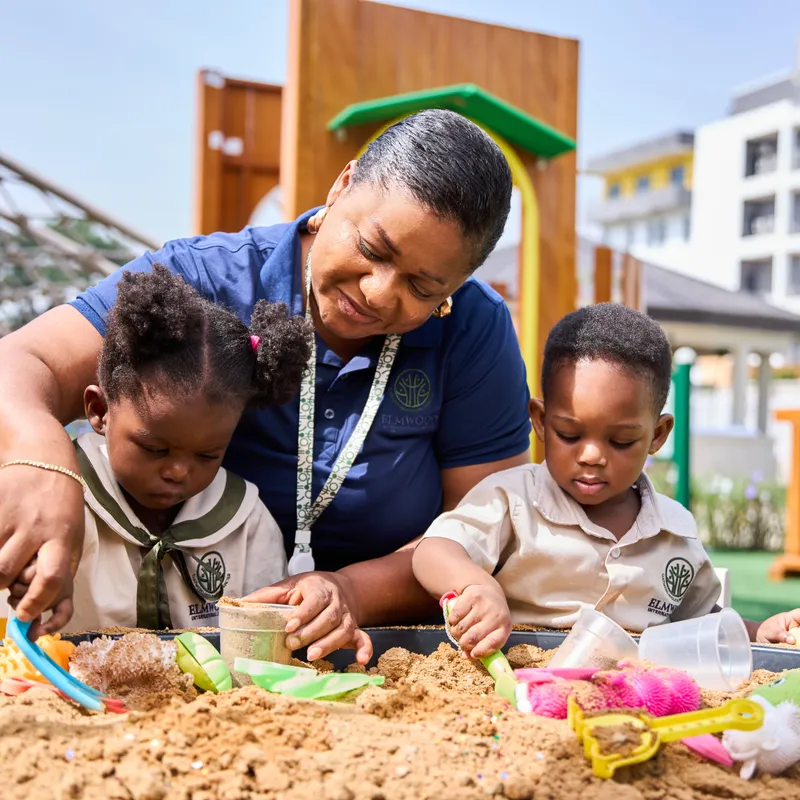 A young Elmwood International School student wearing a black chef's hat whisking ingredients in a large metal bowl with a smudge of flour on her forehead.